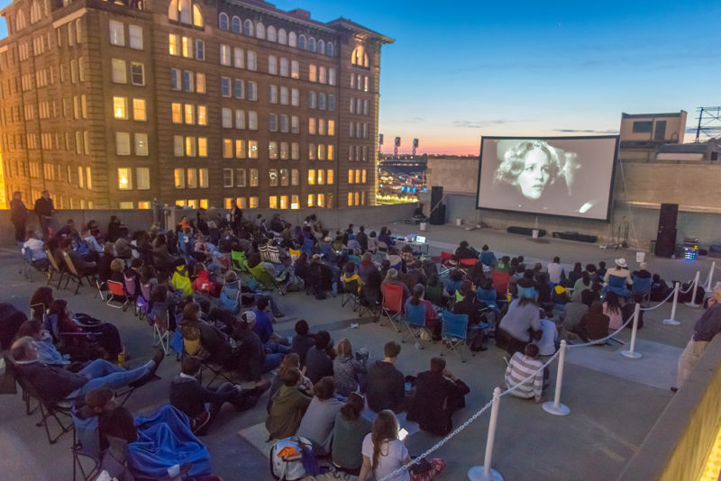 A large group of people sitting in lawn chairs and on blankets on the roof of a concrete parking garage at dusk. There is a movie screen on the roof playing a black and white movie. Pictured on the movie screen is a woman with long, wavy hair and a man with a black hat looking at her. In the background, there is a large, brown, rectangular building with many windows. A baseball stadium is in the far background. The blue sky is in the top of the photo.