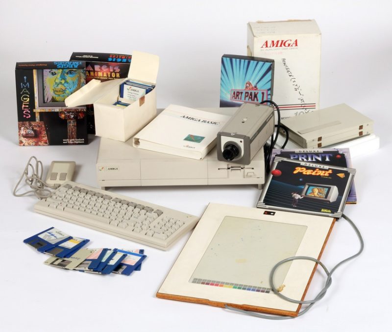 A photograph of an Amiga computer from the 1980s with various other supplies including floppy disks, an art pad, and the packaging for different digital art computer programs.