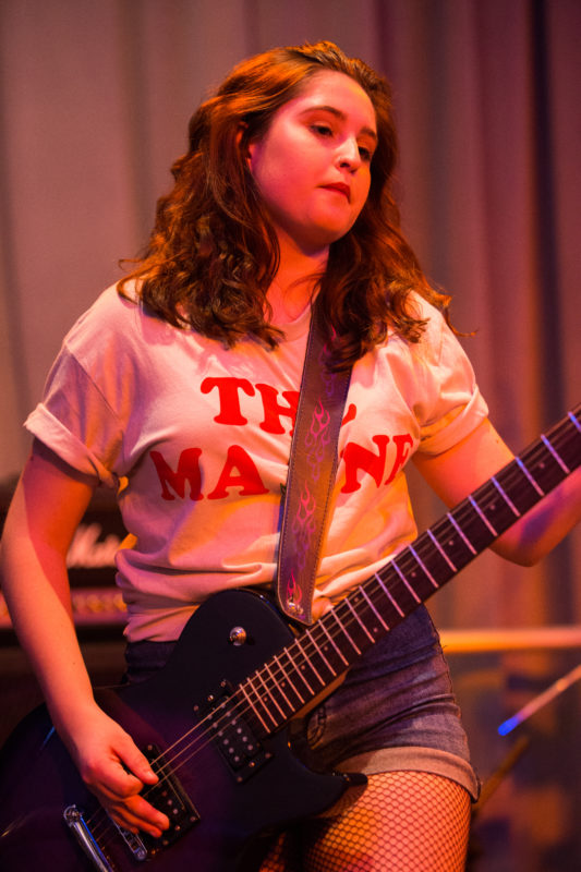 Teen girl with long wavy brown hair a white t-shirt, jean shorts and fish net stockings is playing a blue guitar. The image is cropped so that the bottom of the picture ends above the girls’ knees and slightly above her head.