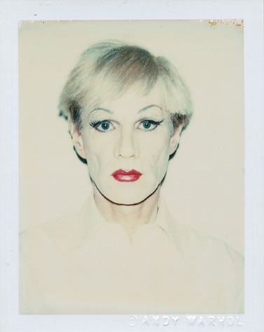 A black and white photograph shows Andy Warhol from the chest up in drag, wearing heavy face powder, black eyeliner, and dark red lipstick. He is facing forward and stares at the camera, with a loose white blouse unbuttoned at the neck.