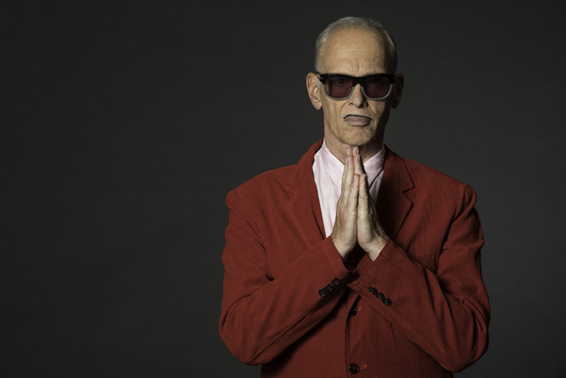 A man with short, thinning gray and white hair is standing facing the camera. He has a thin, black mustache and is wearing sunglasses. His hands are in front of his face pressed together in a praying-type pose. He is wearing a red suit coat and a white shirt.