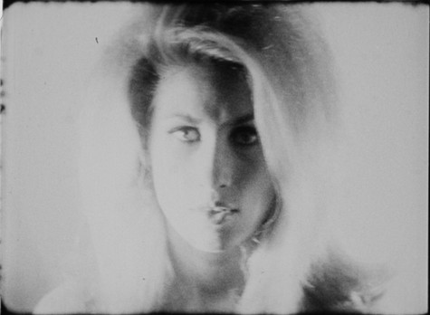 A blonde woman with voluminous hair and heavy eye makeup gazes directly into the camera in this black-and-white film still. The image is blurry and full of light, giving the image a dreamlike quality.