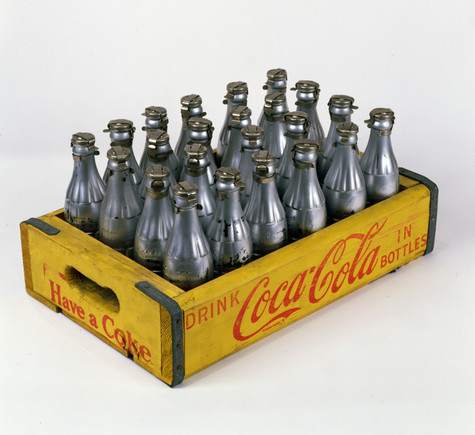 This artwork consists of 23 Coca-Cola bottles spray painted silver and arranged in a silkscreen printed, wooden crate. The crate is painted yellow with lettering printed in red. The slogan on the front panel reads “Have a Coke” while the logo on the right-side panel reads Coca Cola along with the words “drink” and “in bottles”.