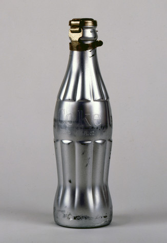 A Coca-cola bottle which has been coated with silver paint sits alone against a gray background. The word Coke can be made out around the middle of the bottle, where the label would be.
