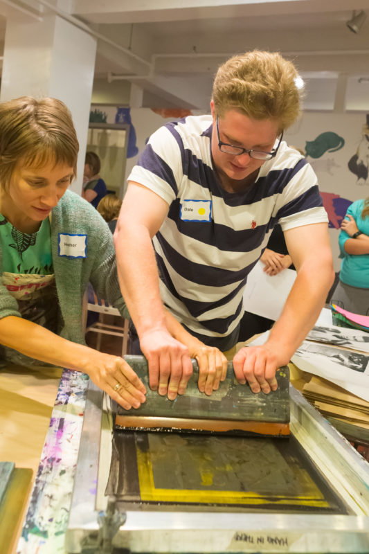 A program participant pulls a squeegee across a silkscreen, creating a print with the assistance of one of The Warhol's Artist Educators.