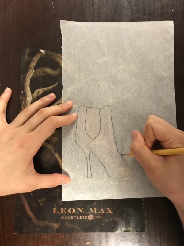 A piece of tracing paper has been placed over a Leon Max magazine advertisement. A pair of hands use a pencil to trace the outline of a shoe featured in the ad.