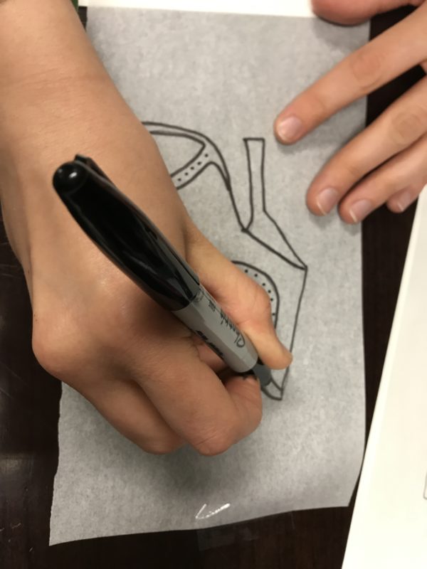 A pair of hands re-traces the outline of a shoe on tracing paper with a black Sharpie marker.