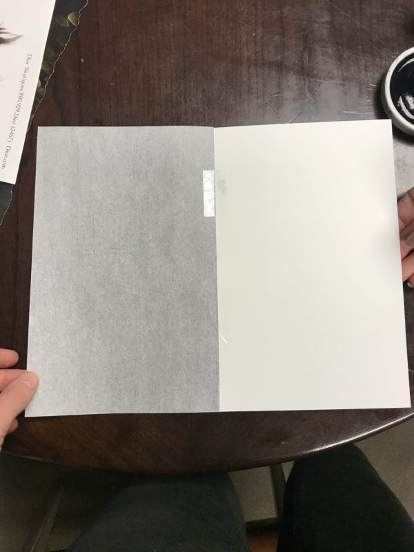A piece of tracing paper attached to a piece of watercolor paper with two pieces of tape functioning as hinges, holding the pieces of paper together.