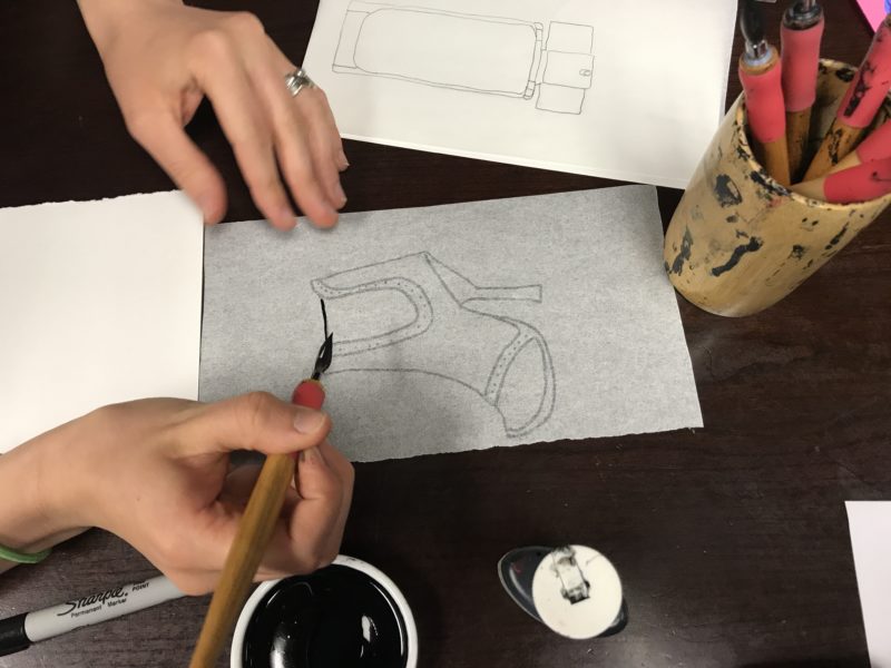 With the tracing paper attached to a piece of watercolor paper, a student uses a paintbrush to apply ink to the image of a shoe that they have drawn on the tracing paper.