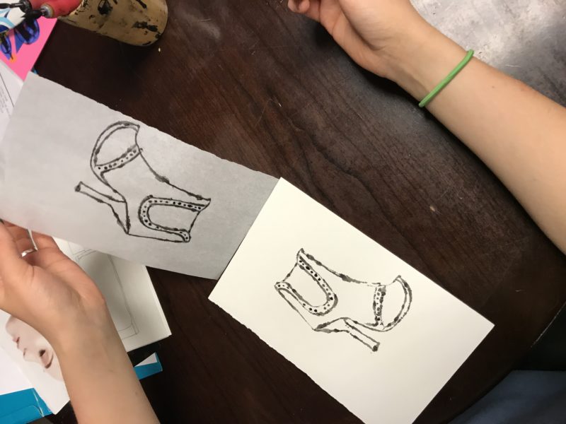 The student pulls away the piece of tracing paper, revealing that the image of a shoe has been transferred to the watercolor paper.