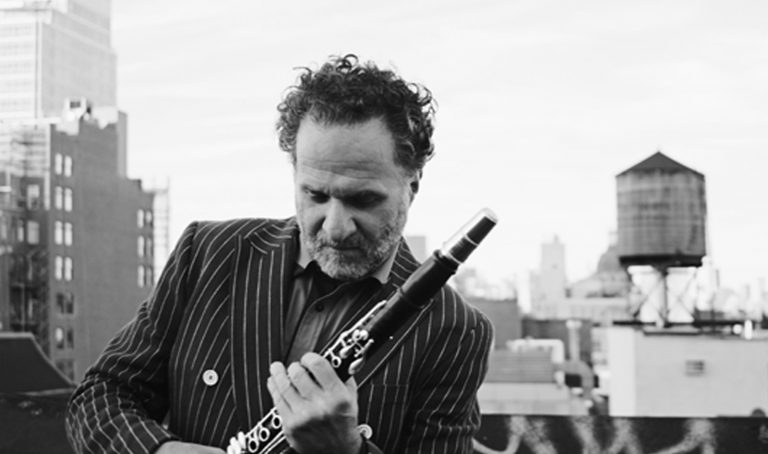A black and white photograph of a man standing on the rooftop of a building with buildings and a watertower behind him. He has short salt and pepper colored hair a salt and pepper colored mustache and beard. He is wearing a black suit coat with white pinstripes and white buttons. He is holding a clarinet instrument in his hands in front of him and is looking down at the instrument.