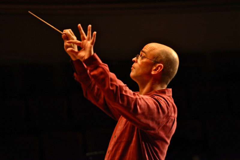 A man who is bald and wearing glasses and a red, long-sleeve, button-down shirt is conducting an orchestra. He has both of his hands raised up in front of him and is holding a conductor's stick in his right hand.