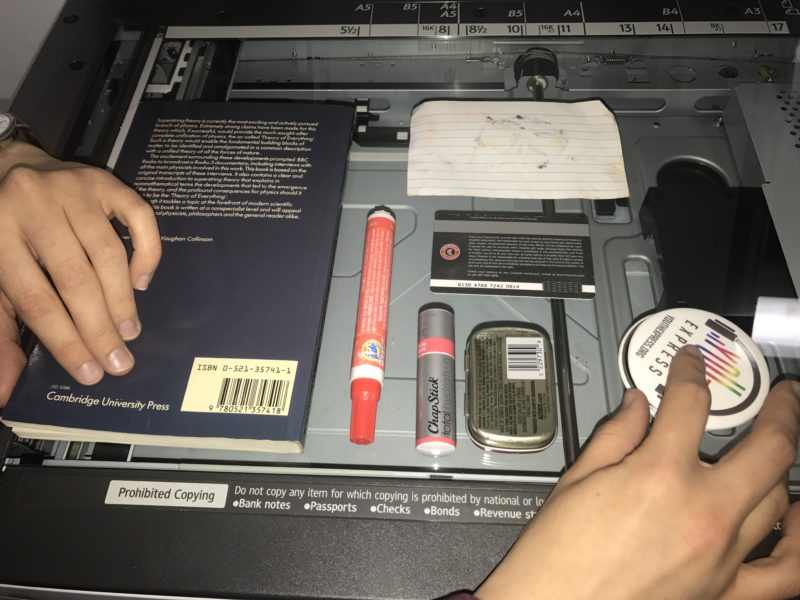 A series of objects is arranged on the glass surface of a photocopier. At left, a student’s left hand adjusts a book, which is face down without a visible title. In the center of the glass surface, a worn 3″ by 5″ index card, a gift card, a small tin of mints, a clearly marked ChapStick, and a bright-red Tide-to-Go pen are arranged to fit the space. These objects are facedown on the glass for copying. On the right, the student’s right hand partially covers a pin with the words “Youth Express” inside an image of headphones.