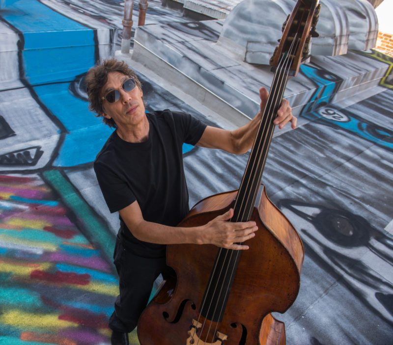 A man with short, brown hair in a black shirt and black pants, wearing dark sunglasses, holds a string bass instrument on top of a roof that is painted on in various colors.