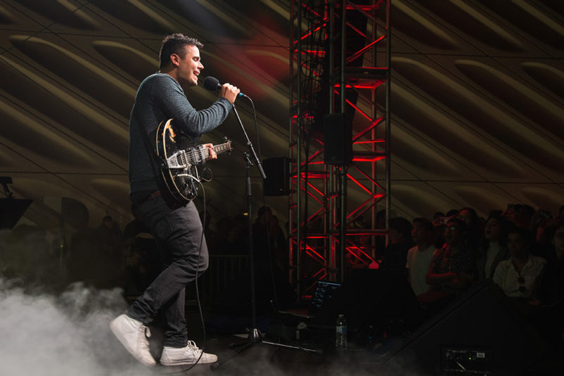 A man with short, brown hair in a gray shirt, gray pants, and white sneakers has a black guitar with white accent lines on the edges on his shoulder. One hand is on the neck of the guitar while the other is holding a microphone on a microphone stand. He is singing into the microphone in front of a crowd of people. There is scaffolding in the background that is lit up in red. There is also fog around his feet.