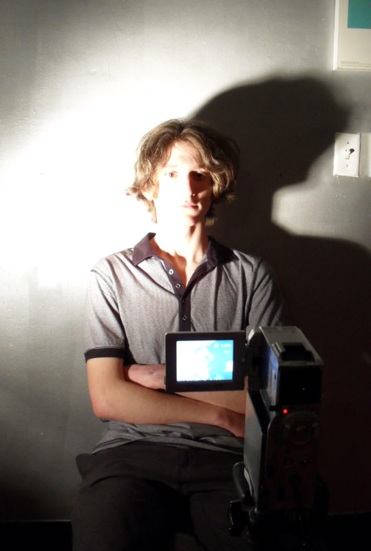 A young man sits with his arms crossed over his abdomen in front of a video camera. He has a slim build and shaggy light brown hair, and is wearing a short-sleeved gray polo shirt and gray trousers. A bright light from the left is shining on his face, partially obscuring his facial features while creating strong shadows to the right. A light switch can be seen on the wall behind him to the right of his head.