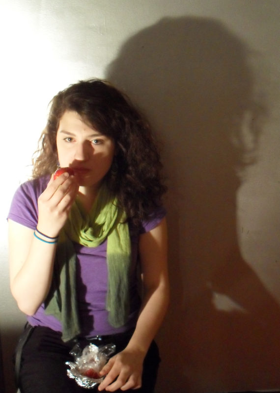 A teenage girl with curly, brown hair and brown eyes sits eating an apple with her right hand, against a silver wall. A bright light shines on her from the left, casting a shadow of her head and arm on the wall to the right.