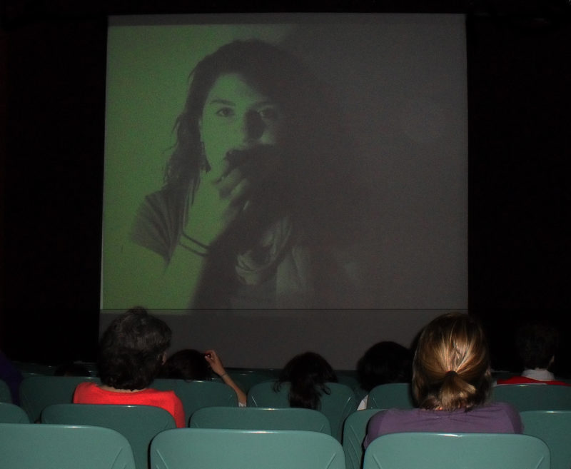 A group of people watch a projection of a young woman on a theater screen.