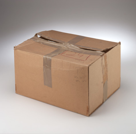 A worn, brown cardboard box sits against a white background. It had been sealed with silver duct tape, but it has been opened. The number “21” is written in red pen on top of the box. There is also minimal additional writing on the side of the box in pencil.