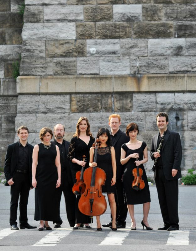 A group of either people, four men and four women, stand facing the camera. The men are wearing black shirts, black sportcoats and black pants. The women are wearing black dresses. One woman in the middle of the photo is holding a stand-up string bass. The women to her left and right are holding violins. The group is standing on a street in front of a large, stone building.