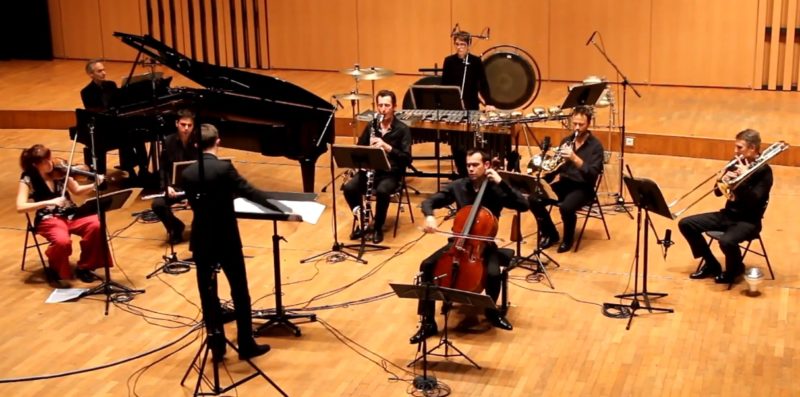 An ensemble of nine people is playing a song on a stage. The ensemble is comprised of eight men and one woman. There is a conductor to the left of the frame facing the rest of the ensemble. Everyone is wearing black shirts and pant, except for the woman who has a black and white blouse and red pants. She is playing a violin. One man on the left is playing a piano. and the others are playing various string, percussion and horn instruments.