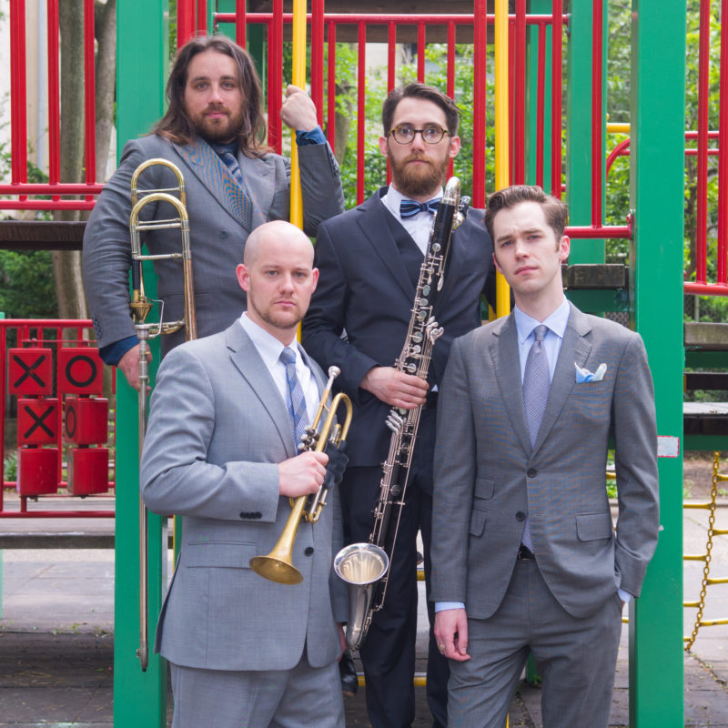 A group of four men are standing, facing the camera in a playground. They are all wearing suits in different shades of gray with dress shirts and ties. Two men are standing in front of the other two men. The man in the foreground on the left is bald with a mustache and facial hair. He is holding a trumpet in his right hand. The man in the foreground on the right has short, brown hair and his left hand is in his pants pocket. The man in the background on the left has long, brown hair that goes to his shoulders and a mustache and beard. He is holding a trombone. The man in the background on the right has short, brown hair, mustache, beard and glasses. He is holding a bass clarinet.