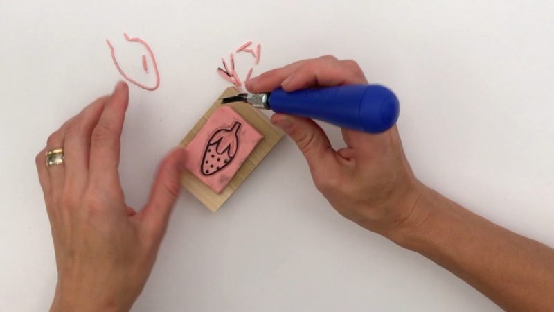 A student's hands hold a blue-handled linoleum carving knife as she cuts the image of the strawberry out of the stamp's pink surface. Linoleum shavings are scattered around the student.