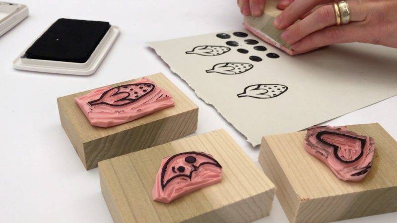 A student is pressing a stamp to a white piece of paper which has already been stamped with several strawberries and dots in black ink. An ink pad, a strawberry stamp, a moon stamp, and a heart stamp are visible in the workstation around the student.