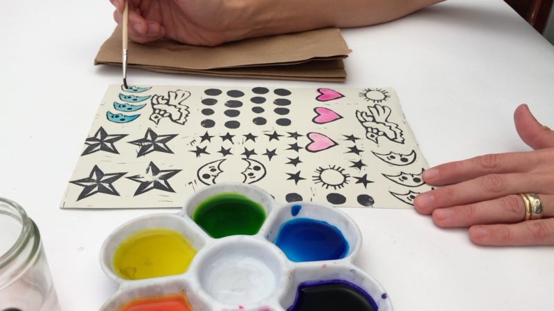 With several different designs stamped on a piece of paper, the student uses watercolor paints to fill in some of the images. Some of the hearts have been filled in pink, and the student is painting the crescent moon designs blue.