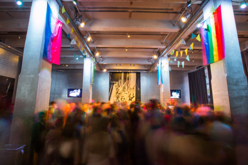 An excited crowd of young people fill the high-ceilinged lobby of The Andy Warhol Museum. They are dressed up in suits and gowns. The silver pillars of the room have decorative flags strung between them. On each pillar there is a flag representing gay, trans, genderqueer, and bi pride. The crowd is blurry, as if they are dancing.