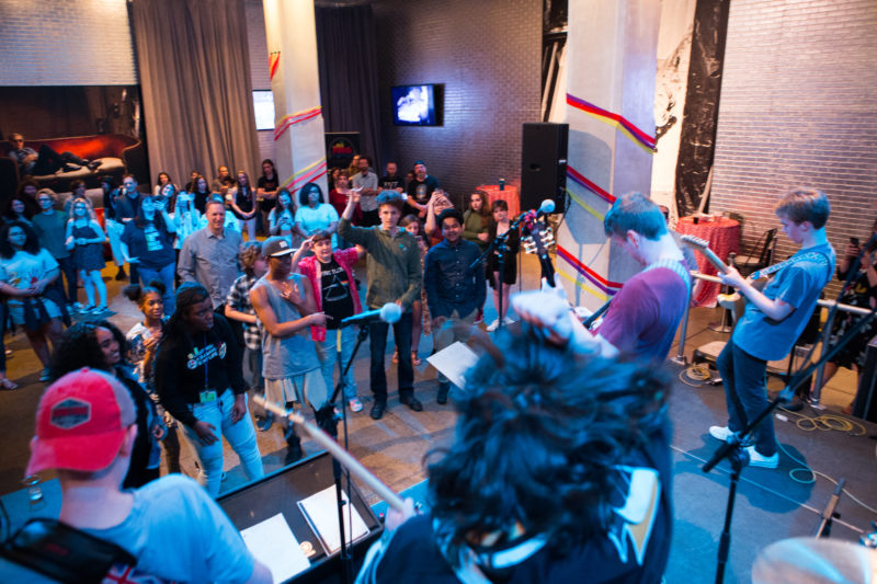 A male teen band performs on a stage in front of crowd in The Warhol entrance space.