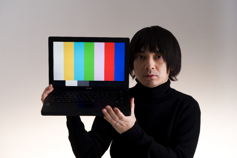 A man with black hair and wearing a black turtleneck sweater is holding a black Apple MacBook laptop with both of his hands. There is a colorbar on the screen of the laptop.