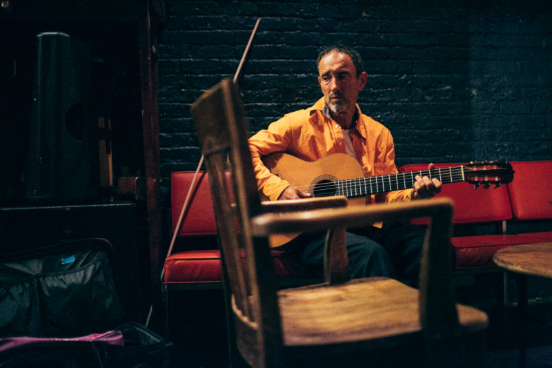 Sitting on a red leather couch, a man is centered in the photograph. He is holding an acoustic guitar that blends in with his mustard yellow shirt. At the foreground of the image lies a wooden chair that is slightly out of focus, creating a grainy and blurred silhouette.
