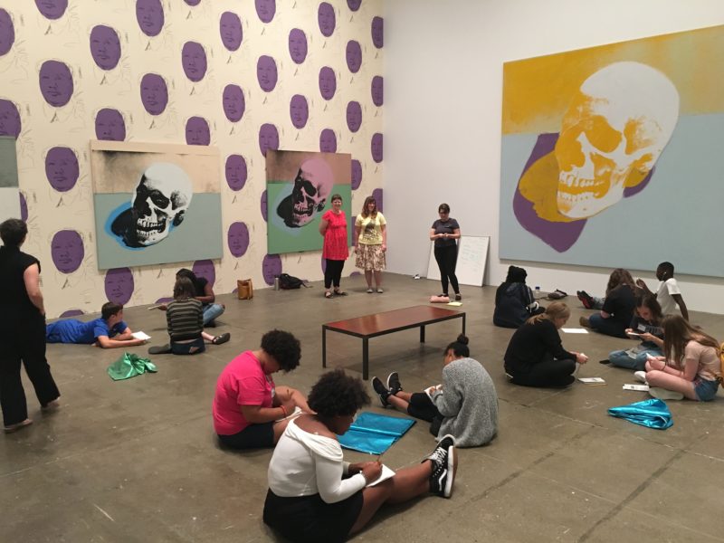 A large group of teens is sitting on the floor and drawing in a gallery with Andy Warhol's skull screen prints. There are three adults standing in the corner.