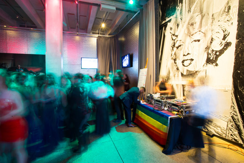 A group of teens dancing in front of a DJ booth with a rainbow flag on it.