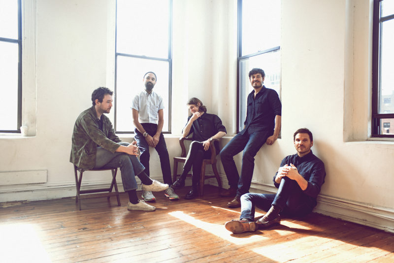 A group of five men in the corner of a room with two, large, windows on each side. Two men sit on chairs, two are sitting on a window ledge, one is sitting on the hardwood floor. Three of them are looking and smiling at the camera. The other two are looking off to the sides.