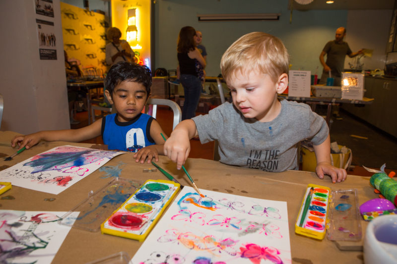 Two toddlers are sitting at a table that has two pieces of paper with butterflies screen printed on them. The toddler on the right is using a paintbrush to paint onto the paper in front of him. The toddler on the left is watching the other toddler paint while holding a paintbrush in his right hand.