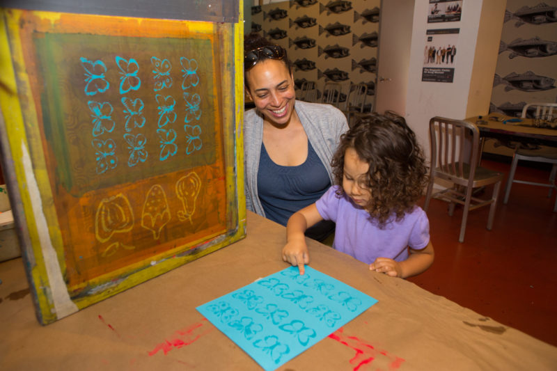 A toddler points at a blue piece of paper with screenprinted butterflies on it with her right hand on a table. In the upper-left-hand corner of the photograph there is a yellowish screen with multiple butterfly and fruit images resting on the table.