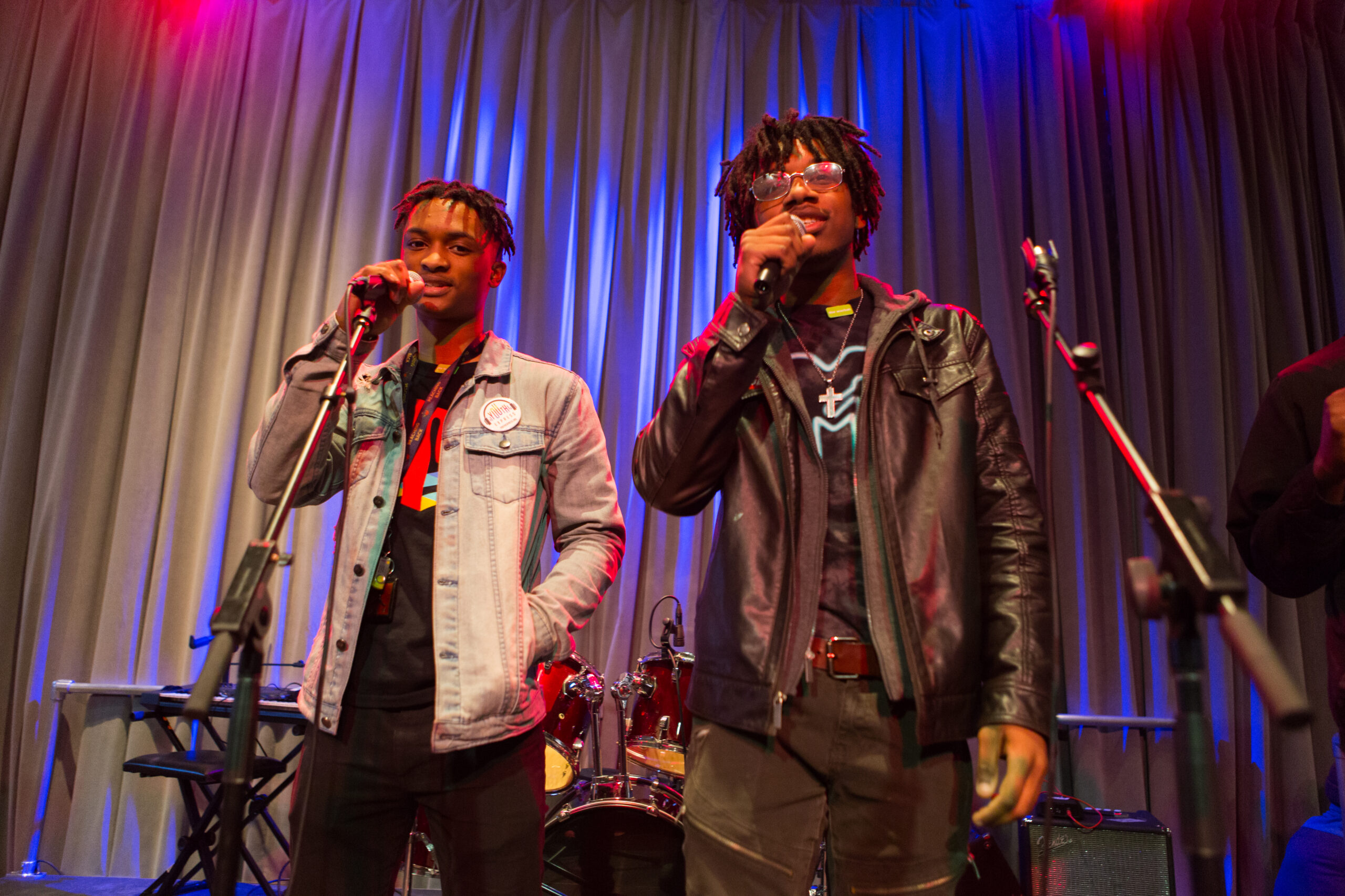 Two young male performers on a stage, sing into microphones.