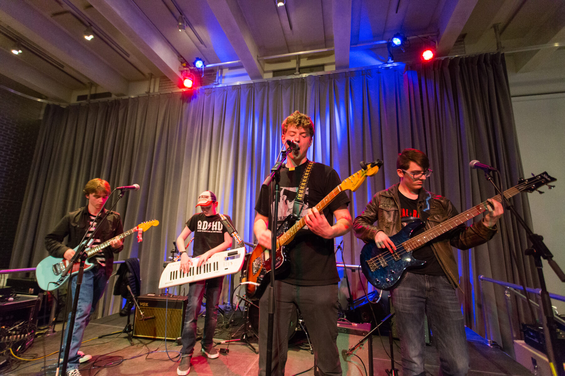 Four young male musicians perform on stage. Each one holding a different instrument - guitar, keytar, guitar and bass.