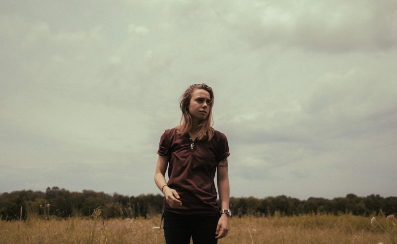 A woman with shoulder length blonde hair is standing in a field. She is looking towards the right side of the frame. She is wearing a maroon shirt, black pants, and a watch on her left wrist.