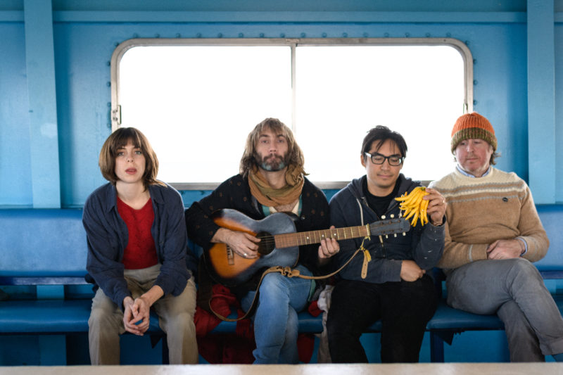 A group of four people sitting in a transit vehicle of some sort. They are sitting on one long, blue seat that's attached to the blue walls. There is a giant window behind them. On the left is a woman with short, brown hair wearing a red sweater, blue coat and khaki pants. She's leaning forward and looking at the camera. The man next to her has long, blonde hair and brown and gray facial hair. He is wearing a brown scarf, black sweater and blue jeans. He's holding a black and brown acoustic guitar. The man to the right of him has short, black hair and is wearing black-framed glasses. He's wearing a gray sweatshirt and black pants and is holding a group of yellow vegetables of some sort. The man to the right of him has short, blonde hair that is under an orange and brown knit cap he is wearing on his head. He is wearing a brown sweater and gray pants.