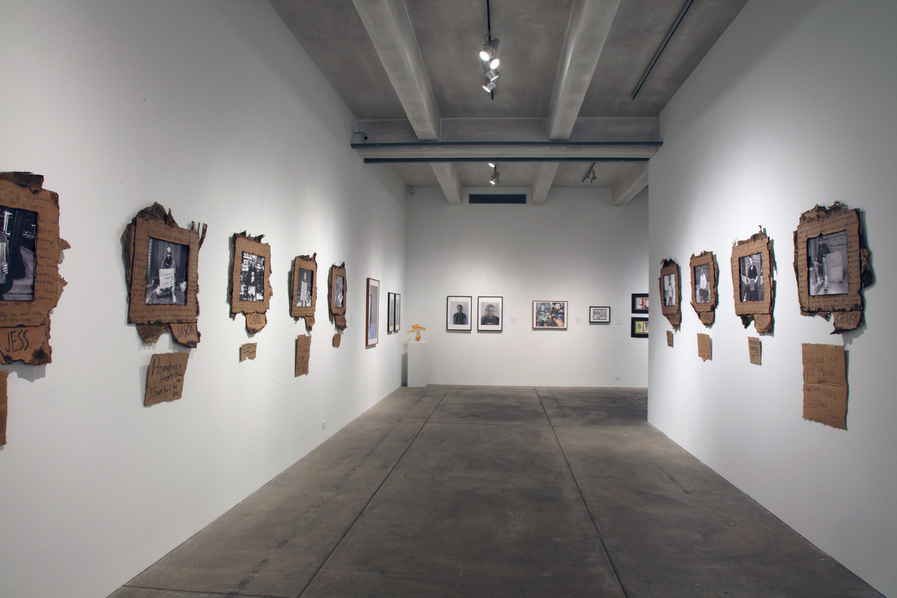 Corridor of a gallery. White walls with black and white photography that is framed in fraying cardboard.