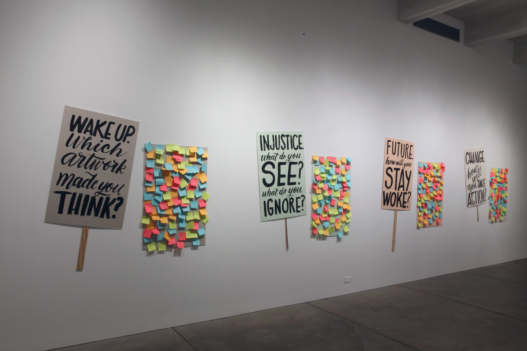 Response prompts styled to resemble protest signs hang on gallery walls adjacent to the corresponded responses, written on sticky notes