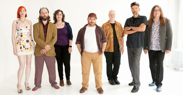 A group of seven people stand in front of a white background looking at the camera. There is one man in the middle of the frame with his hands in his pockets. Three people are standing to his right and three to his left.