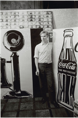A black and white photo of Andy Warhol, wearing dark pants and al light button-down shirt, standing in his study beside two paintings, one of a telephone and one of a coca cola bottle, each approximately as tall as he is.