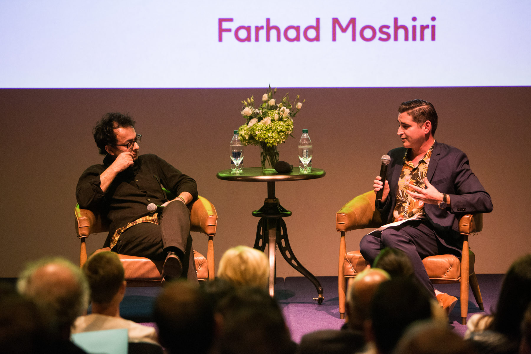 Two men sit on chairs on a stage, engaged in conversation. The screen behind them says Farhad Moshiri.