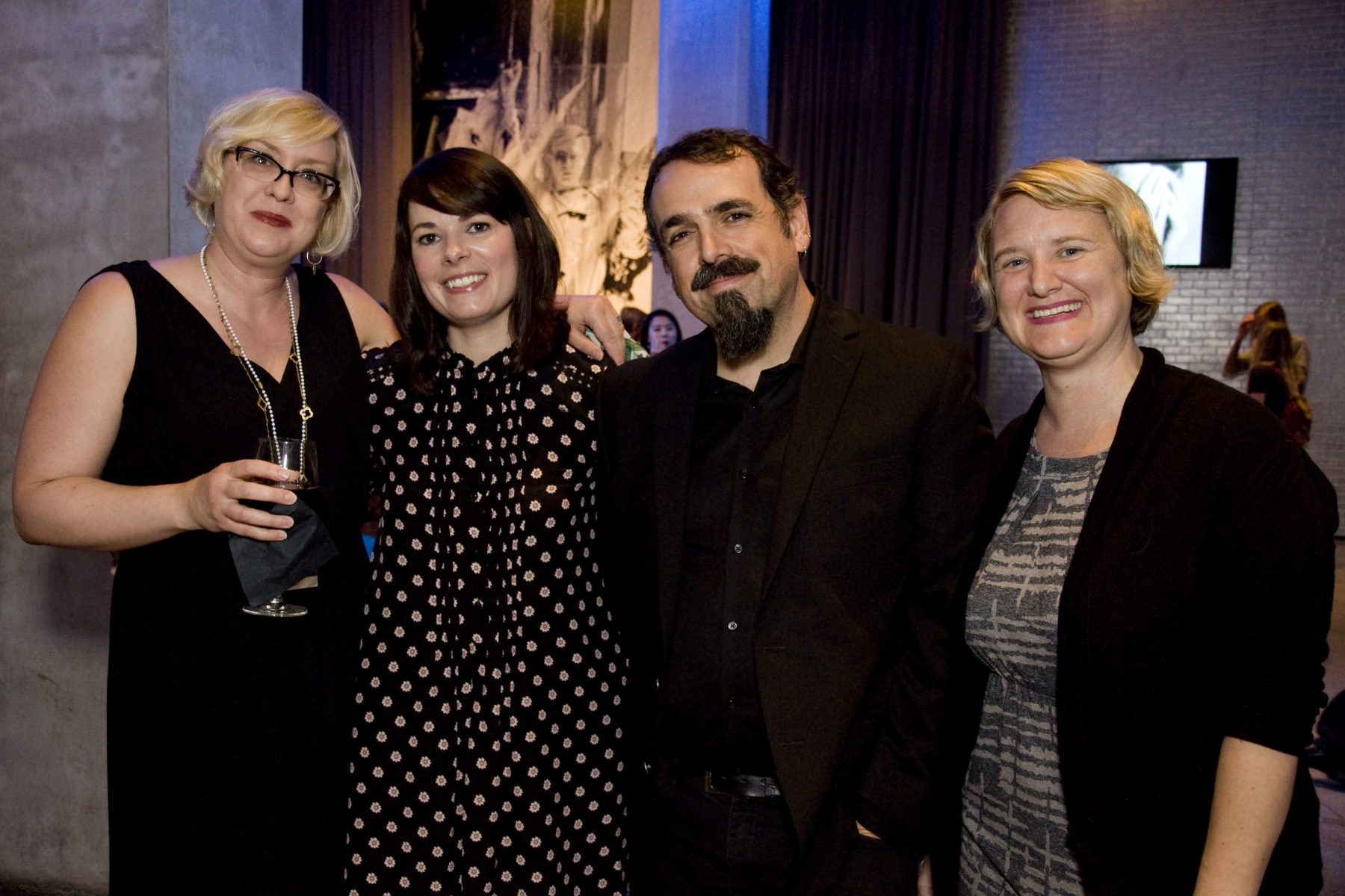Four people dressed in cocktail attire pose for a photo during an exhibition opening. The woman on the far left holds a wine glass.