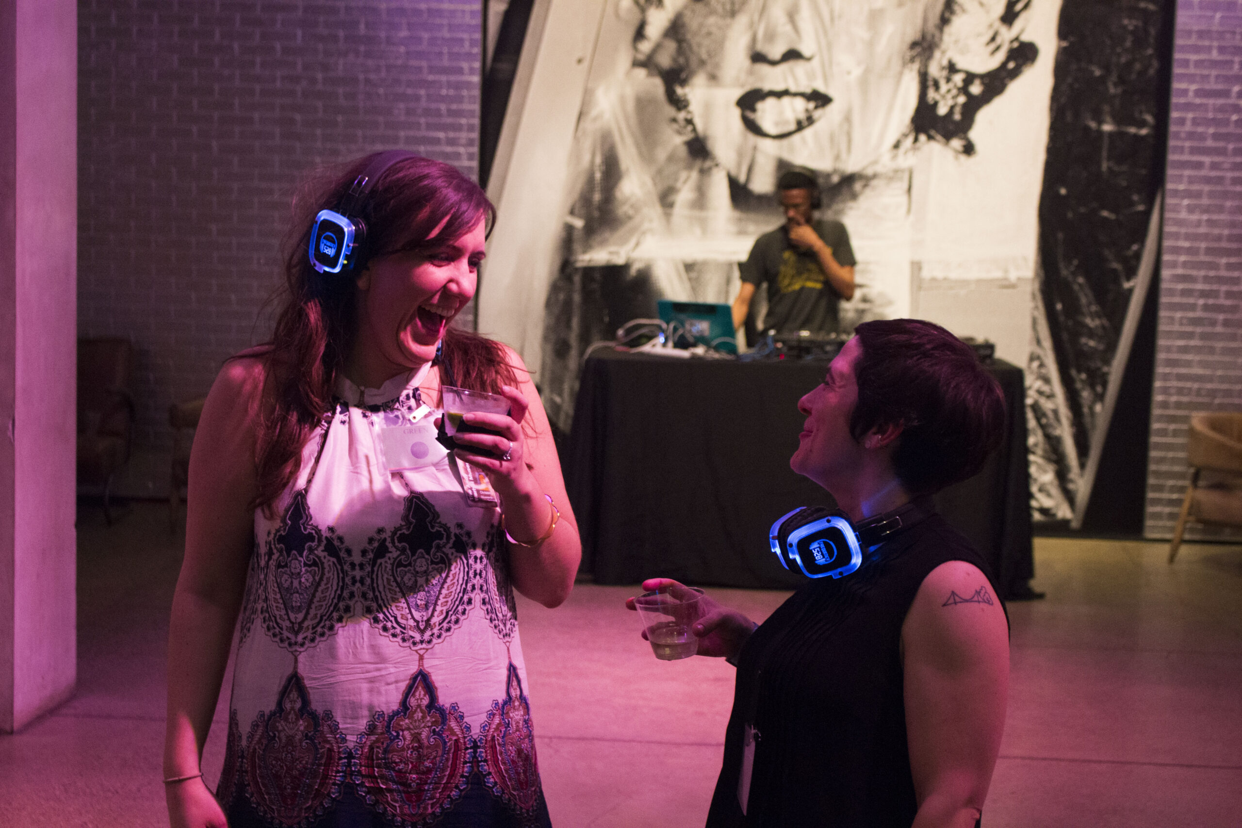 Two women wear noise-canceling headphones that light up bright blue. They are holding cocktails, looking at each other and laughing.