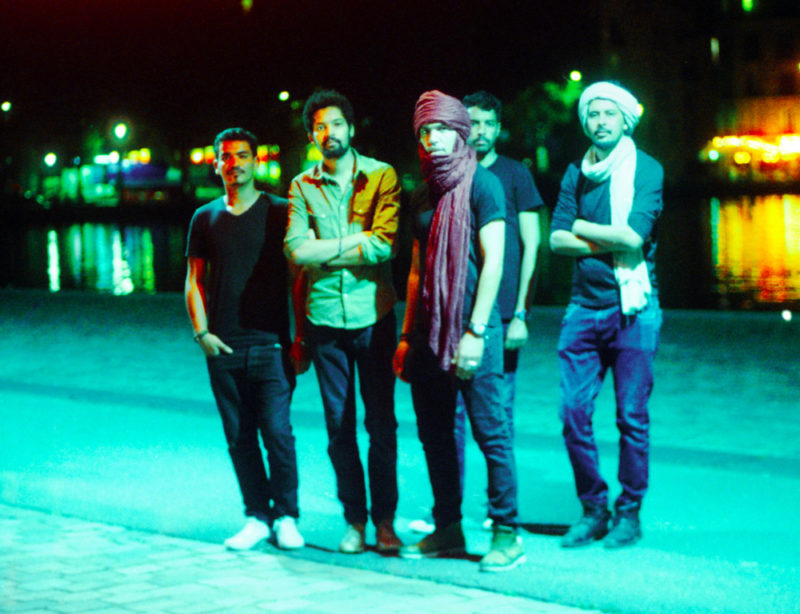 A photograph of four men standing on a street with a river in the background. Three of the men are standing in front of the two others. They are all looking at the camera. Two men have their arms folder and the man on the left has his right hand in his pants pocket.